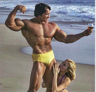 Arnold holding a glass of wine and posing on the beach with a woman on his leg