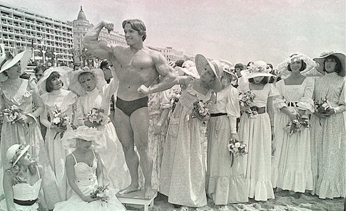 Arnold posing in front of a group of women in white dresses