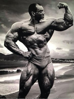 Sergio Oliva on a beach flexing his biceps